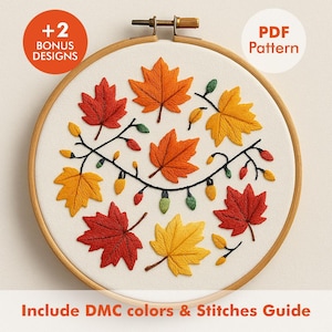 Può includere: Un telaio da ricamo presenta un disegno di foglie autunnali nei toni del rosso, arancione e giallo, con una stringa di luci. Il testo include "+2 Bonus Designs" e "PDF Pattern". Il disegno è ambientato su tessuto color crema, all'interno di un telaio di legno.
