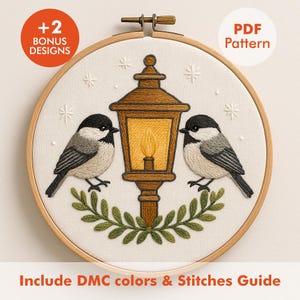 May include: Embroidered hoop art featuring two birds flanking a brown lantern with a glowing yellow light. The design includes green foliage and snowflake accents on a cream background. The text reads "+2 BONUS DESIGNS" and "PDF Pattern".