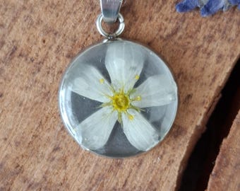 May include: A silver pendant necklace with a clear, round resin charm. The charm contains a white flower with a yellow centre. The necklace chain is also silver. The background is a wooden surface.