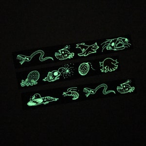 May include: A set of glow-in-the-dark stickers featuring various sea creatures, including a jellyfish, a squid, a fish, and a seahorse. The stickers are black and white and have a whimsical, cartoon-like style.