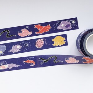May include: A roll of washi tape with a dark blue background and colorful illustrations of deep sea creatures, including a squid, a shrimp, a fish with a lure, and a jellyfish. The tape is on a white surface.