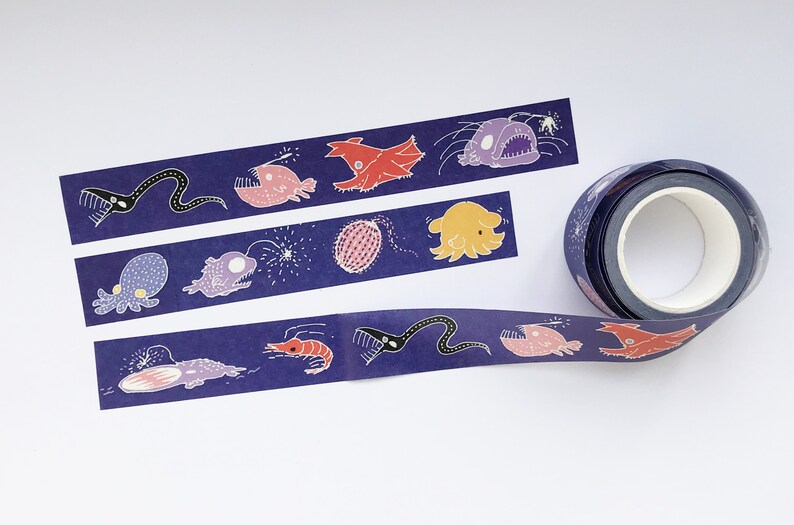 May include: A roll of washi tape with a dark blue background and colorful illustrations of deep sea creatures, including a squid, a shrimp, a fish with a lure, and a jellyfish. The tape is on a white surface.