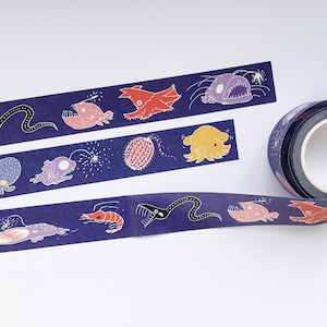 May include: A roll of washi tape with a dark blue background and colorful illustrations of deep sea creatures, including a squid, a shrimp, a fish with a lure, and a jellyfish. The tape is on a white surface.