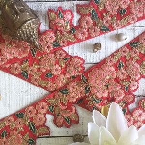 Può includere: Bordura ricamata floreale rossa con accenti dorati e fiori rosa. La bordura è disposta su una superficie di legno bianco con elementi decorativi, tra cui una testa di Buddha in ottone e piccole campane. Ideale per progetti di cucito.