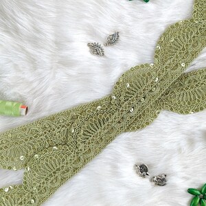 May include: A long strip of olive green lace trim featuring a scalloped edge and intricate floral embroidery accented with small, reflective sequins. The lace is arranged in a cross shape, accompanied by two small spools of green thread.