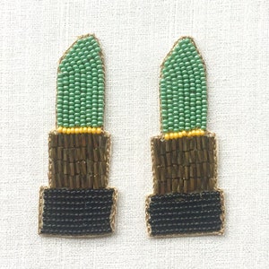 Peut inclure: Une paire de boucles d'oreilles en forme de rouge à lèvres en perles vertes, dorées et noires. Le rouge à lèvres est fait de perles vertes, la base est en perles dorées et le bas est en perles noires.