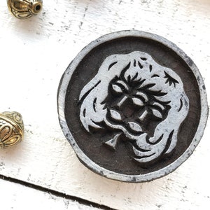 Op de afbeelding: Een ronde zwarte en witte stempel met een gedetailleerd ontwerp van een gezicht met lang haar. De stempel is gemaakt van metaal en heeft een ruwe textuur.