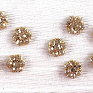 May include: Ten small, gold-colored, flower-shaped embellishments with clear rhinestones. Each embellishment has a circular base and a raised flower design.