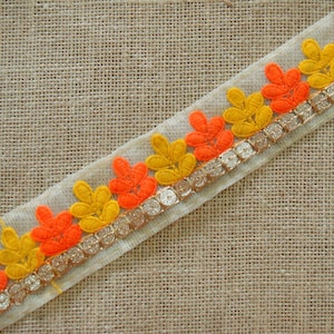 May include: Orange and yellow embroidered trim with a gold glitter accent. The trim is on a beige fabric background.
