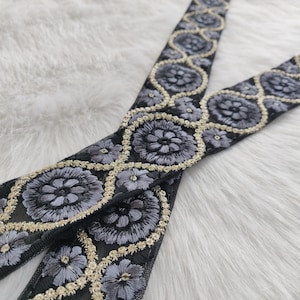 以下が含まれることがあります： 花柄のデザインが施された、2本の黒い刺繍リボンのクローズアップ。花はグレーで、金色の糸で縁取られています。リボンは白くふわふわした表面にあります。