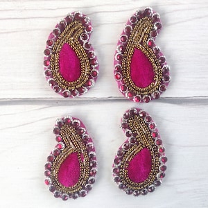 May include: Four pink paisley-shaped fabric embellishments with gold and red beading. The embellishments are on a white wooden surface.