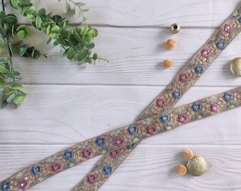 May include: Two strips of decorative ribbon with a floral pattern in blue, pink, and gold. The ribbon is embellished with sequins and sits on a light wood surface. Greenery and decorative gold accents are also present.