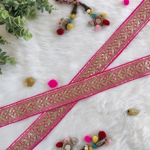 May include: Two strips of vibrant pink ribbon with intricate gold embroidery and sequin detailing. The ribbon features a repeating floral pattern. The background is a soft, white, fluffy surface, with decorative elements like pom-poms and beads.