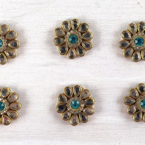 Puede incluir: Seis adornos en forma de flor con base de metal dorado. Cada uno tiene piedras transparentes en forma de lágrima alrededor de una piedra central de color turquesa. Los adornos están dispuestos sobre un fondo de tela blanca.