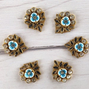 Puede incluir: Cinco adornos dorados con un diseño de flor azul y pedrería transparente. Cada adorno tiene una forma diferente.