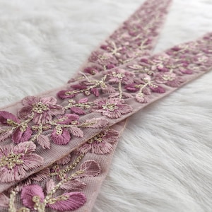 Puede incluir: Primer plano de dos tiras de encaje bordado. El encaje presenta un delicado diseño floral en tonos rosas y morados, con detalles dorados. El encaje se encuentra sobre un fondo suave y blanco texturizado.
