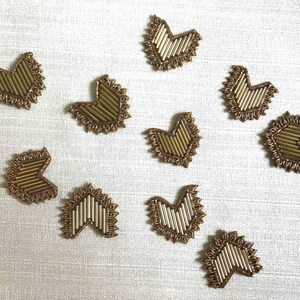 Peut inclure: Dix embellissements en forme de cœur dorés avec un motif en zigzag. Les embellissements sont sur un fond de tissu gris clair.