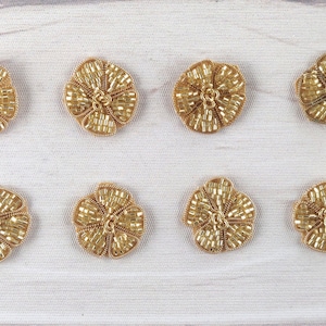 Peut inclure: Huit embellissements en forme de fleur de couleur dorée avec un design en relief texturé. Les embellissements sont disposés en deux rangées de quatre sur un fond blanc.