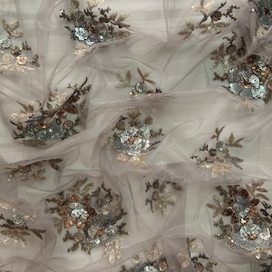 Könnte beinhalten: Nahaufnahme eines durchsichtigen, hellrosa Stoffes mit zarter Blumenstickerei und Pailletten in Gold- und Silbertönen.