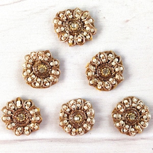 May include: Six gold-colored fabric flower embellishments with pearl accents and a rhinestone center. The embellishments are arranged in a circle on a white surface.
