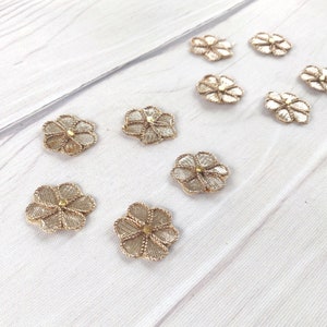 May include: Ten gold-colored flower-shaped embellishments with a gold bead in the center. The embellishments are on a white wooden surface.