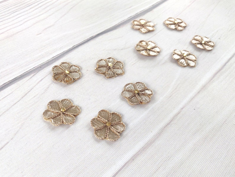 May include: Ten gold-colored flower-shaped embellishments with a gold bead in the center. The embellishments are on a white wooden surface.
