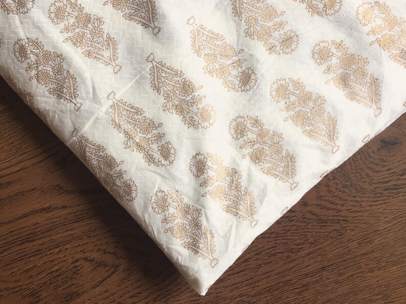 May include: White fabric with a gold floral pattern. The fabric is folded in half and the pattern is visible on both sides.