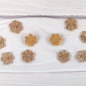 May include: Eight gold-colored flower-shaped buttons with a raised center. The buttons are arranged in two rows of four on a white wooden surface.
