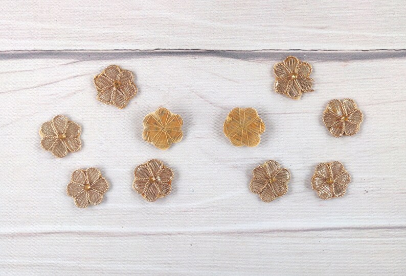 May include: Eight gold-colored flower-shaped buttons with a raised center. The buttons are arranged in two rows of four on a white wooden surface.