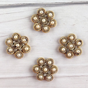 Op de afbeelding: Vier goudkleurige, bloemvormige versieringen met witte parelmoerachtige accenten. Elke versiering heeft een cirkelvormig midden met kleinere cirkels eromheen.