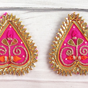 Könnte beinhalten: Zwei rosafarbene herzförmige Stoffverzierungen mit goldener und orangefarbener Stickerei. Die Verzierungen haben einen gewellten Rand und sind mit einem floralen Muster verziert.