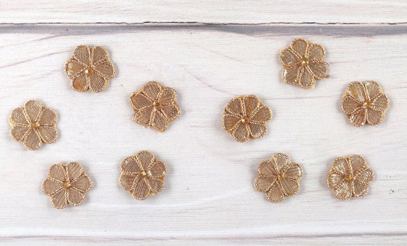 May include: Ten gold-colored metal flower-shaped embellishments on a white wooden surface.