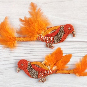 May include: Two orange and red embroidered bird appliques with gold accents and feathers. The birds are facing right and have a long tail of feathers.