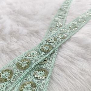 Puede incluir: Primer plano de dos tiras de encaje decorativo. El encaje es de un color verde pálido suave con patrones florales bordados y lentejuelas plateadas. El encaje se encuentra sobre un fondo blanco y esponjoso.