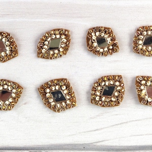 Peut inclure: Dix embellissements en forme de diamant de couleur dorée avec des centres en miroir. Chaque embellissement est entouré de perles blanches et de perles dorées.