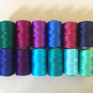 Op de afbeelding: Een set van tien spoelen garen in verschillende tinten blauw, paars en groen. De spoelen zijn in een rij gerangschikt, met de kleuren die van donker naar licht overgaan.