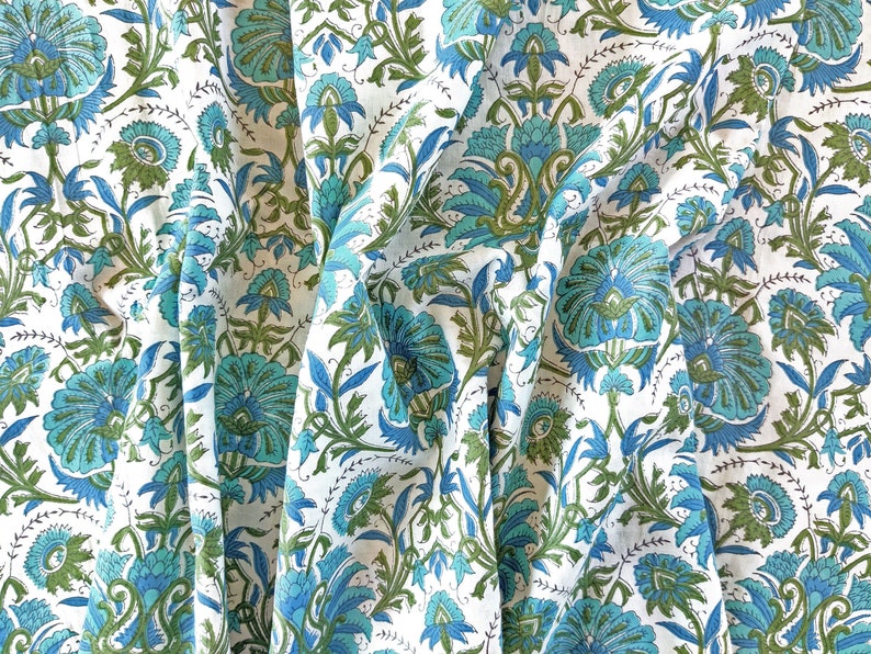 May include: White fabric with a blue and green floral pattern. The fabric is folded and draped, showing the intricate details of the design.