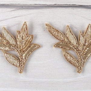 Könnte beinhalten: Zwei goldfarbene, gestickte Blumenapplikationen mit einem Blattdesign. Die Applikationen sind aus Metallic-Garn gefertigt und haben ein zartes, filigranes Design.