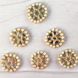 Puede incluir: Seis adornos redondos decorativos. Cada uno tiene un marco de metal dorado con un centro espejado y un borde de pequeñas cuentas blancas con forma de perlas. Los adornos están dispuestos sobre una superficie clara con vetas de madera.