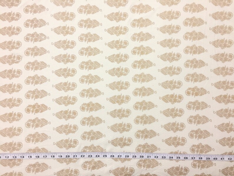 May include: White fabric with a repeating pattern of beige stylized floral sprigs. The fabric has a ruler with inch markings at the bottom.