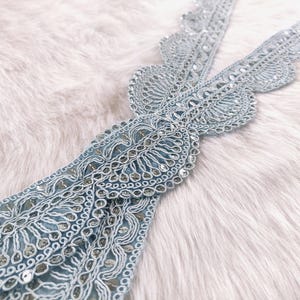 May include: A pale blue lace trim featuring detailed embroidery and shimmering sequins. The trim is displayed on a soft, white faux fur background. This decorative trim is suitable for sewing and craft projects. The trim's design includes floral and geometric patterns.
