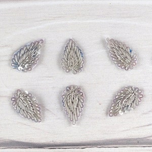 May include: Ten silver leaf-shaped embellishments with a beaded design on a white background.