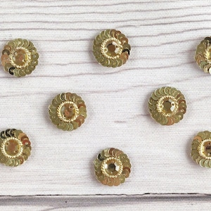 May include: Ten gold round sequins with a raised center, arranged in a scattered pattern on a light wood surface.