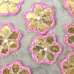 May include: A collection of seven pink and gold flower embellishments with a sparkly finish. The flowers are made of fabric and have a pink border.
