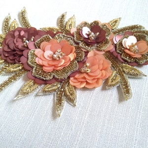 Peut inclure: Une applique florale avec trois fleurs en sequins pêche et marron et des perles dorées. L'applique est entourée de feuilles dorées.