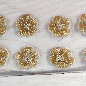 Peut inclure: Huit embellissements floraux en perles dorées avec un éclat métallique. Les fleurs sont disposées en deux rangées de quatre sur une surface blanche.