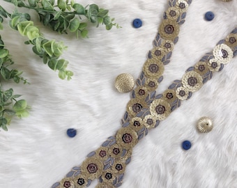 May include: Two strips of decorative trim with gold sequins in a floral pattern, accented with blue and gold embellishments. Green leafy sprigs and small blue and gold buttons are scattered around the trim on a white, fluffy surface.