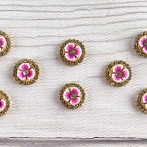 May include: Ten round pink and white flower embellishments with gold trim and clear rhinestone centers. The embellishments are arranged in two rows on a white wooden surface.