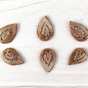 Peut inclure: Dix embellissements en forme de feuille dorés avec un motif en relief au centre. Les embellissements sont disposés en deux rangées de cinq sur une surface blanche.