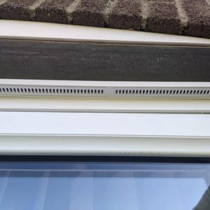 May include: A white window frame with a white vent above the window pane. The vent has a series of small holes for air circulation.